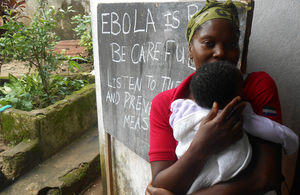 Lady holding a baby in front of a blackboard with information on Ebola prevention