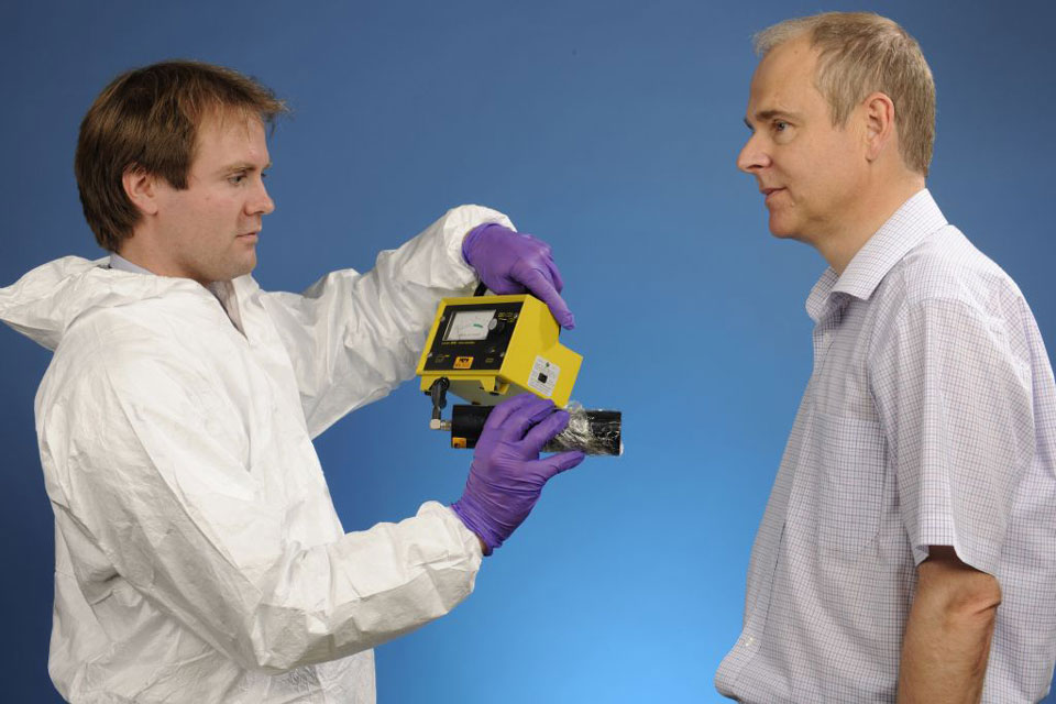 A lab technitian wearing a white lab suit and purple nitrile gloves holding a handheld detector 30 cm away from a man being monitored
