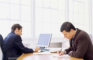 two men at desk with papers and computer
