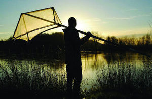 A fisherman with a net suitable for elver fishing standing by a river bank, backlit by the sun