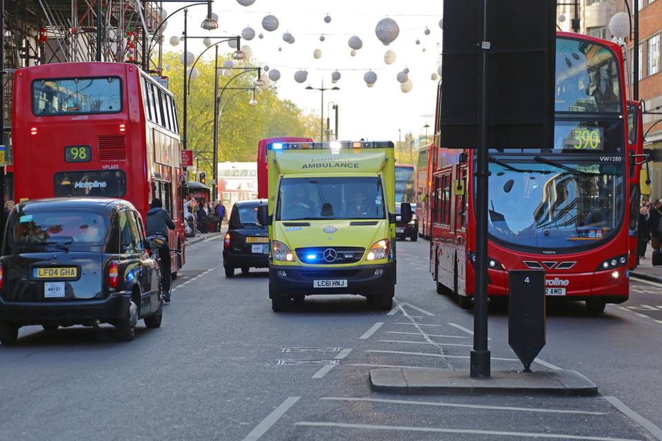 Ambulance on 999 call in London traffic between two red double-decker buses.