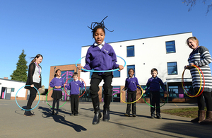 Children playing with hula hoops in front of school watched by teachers against a blue sky.