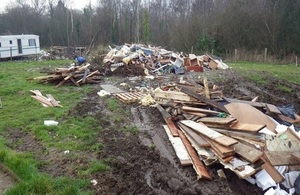 Picture shows piles of wood and building waste in a field, with a caravan in the background
