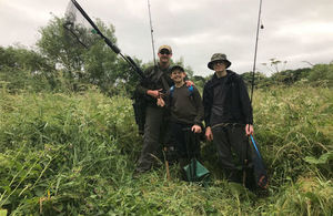 Three anglers standing in tall grass holding their fishing rods and a net