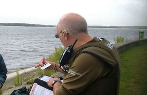 An Environment Agency fisheries enforcement officer standing next to water and viewed from behind checking a fishing licence