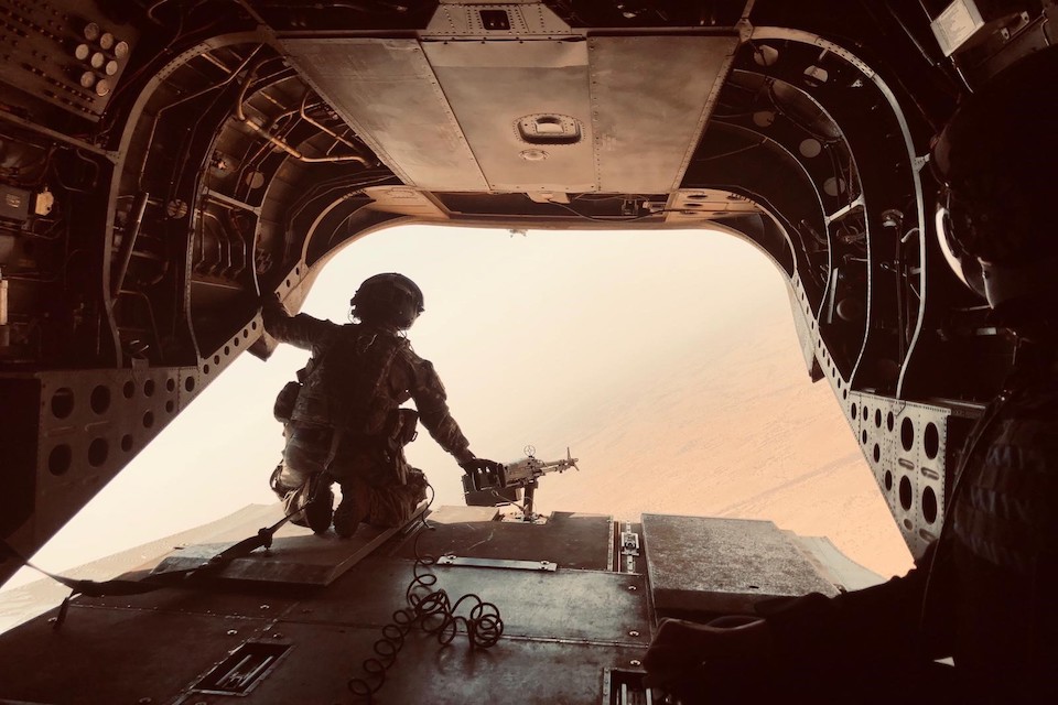 Interior of an aircraft: a soldier in desert camo gear looks out of the back of an aircraft in mid-flight. A desert landscape can been seen below.