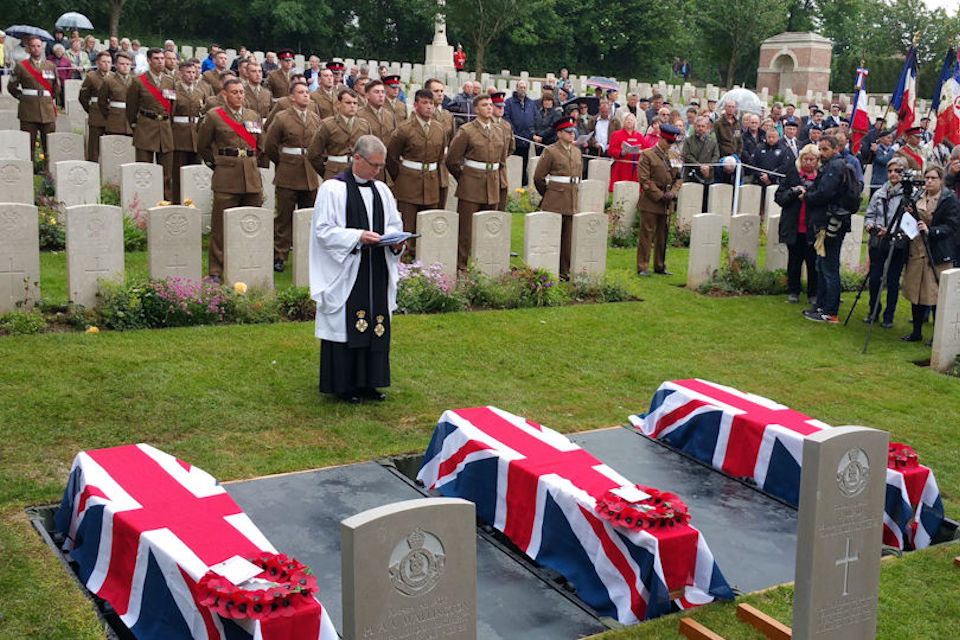 Military funeral service in progress with three coffins on the ground surrounded by military personnel in full uniform. 