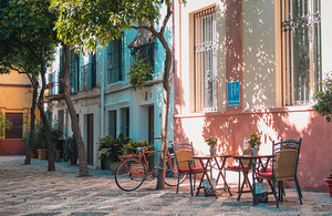 A European street scene with colourful building fronts, trees, table and chairs and a bicycle.