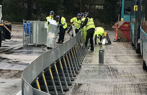 Picture shows workers in yellow jackets assembling metal barriers in a concreted area with buses parked to the right