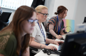 Three women sitting by computers with headphones on dealing with telephone calls