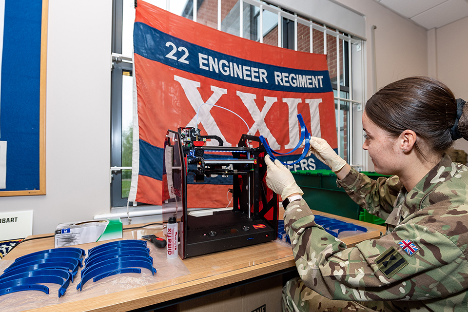 A woman in British Army uniform operates 3D printing machinery, with a row of printed objects beside her. 