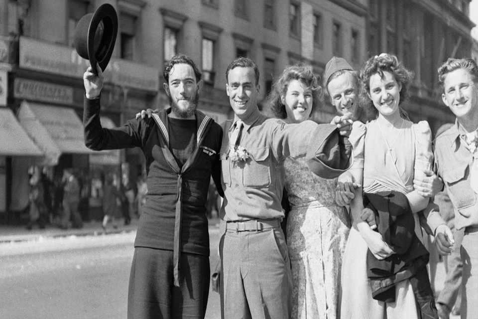 A New Zealand sailor raises his bowler hat in the sunshine after hearing of the Victory in Japan with several American soldiers and English civilian women on the Strand in London.