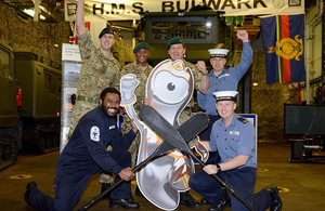 Members of HMS Bulwark's ship's company surround a cut-out of an Olympic mascot to cheer on Senior Service Olympian Lieutenant Pete Reed