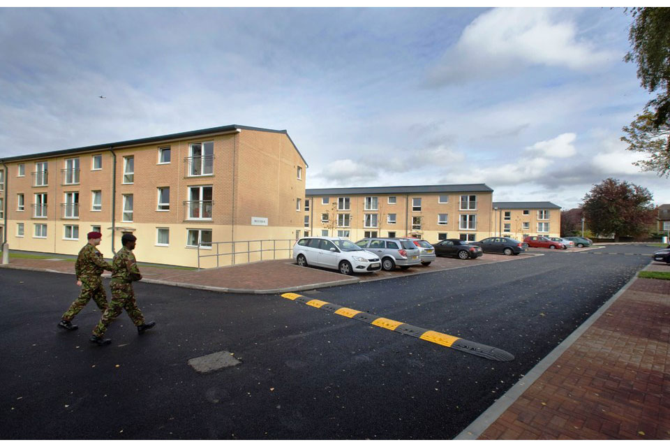 Example of service family accommodation showing 2 military personnel walking past 3 blocks of housing, all of which are 3 storeys high. There is off road car parking and speed bumps.
