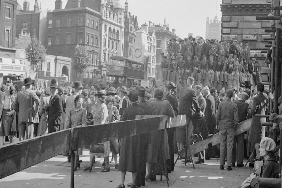 Crowds of people wait for news at the junction of Whitehall with Downing Street. The crowd consists of service personnel and civilians alike, all waiting for confirmation from the Prime Minister that the war in Japan is over. 