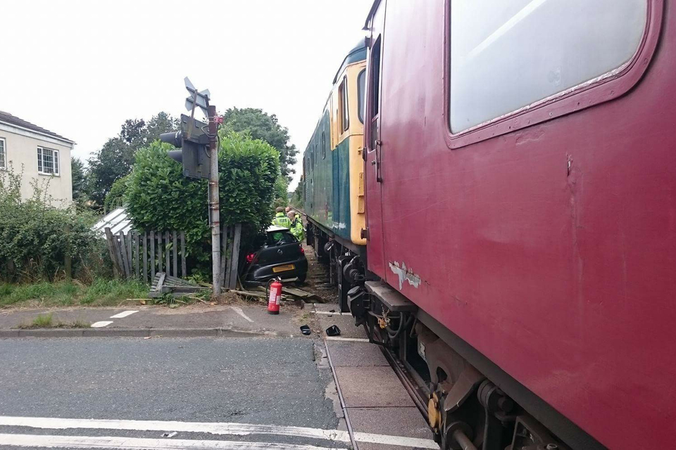 A  locomotive and a burgundy railway carriage stopped on the level crossing at Yafforth. Members of the emergency services are standing near a small hatchback which is in a hedge surrounded by damaged fencing. (image courtesy of the Wensleydale Railway) 