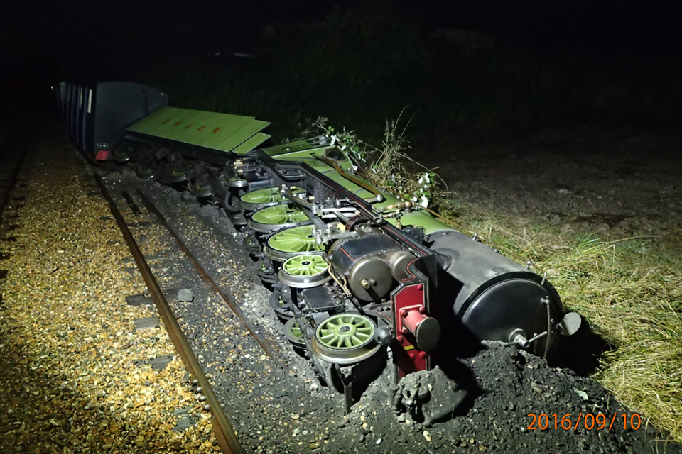 A night time view light by floodlights showing Romney Hythe and Dymchurch locomotive Number 1 'Green Goddess' lying on its left hand side after the accident. The locomotive is partly buried in the black ballast which has piled up around it.