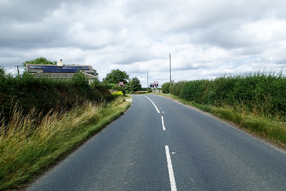 The approach to Yafforth level crossing from the south, showing the road layout around 65 metres from the crossing. The level crossing lights can be seen. A large white house is just to the left of the crossing.