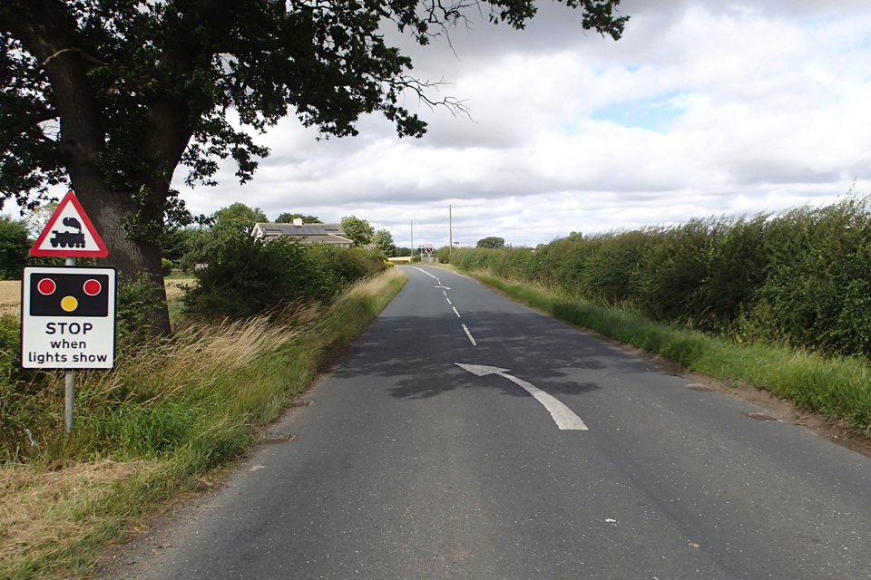 The road on the approach to Yafforth level crossing from the south. Trees and hedges line each side of the road with shadows across the road. A warning sign telling road users to stop when the lights show is on the left hand side.