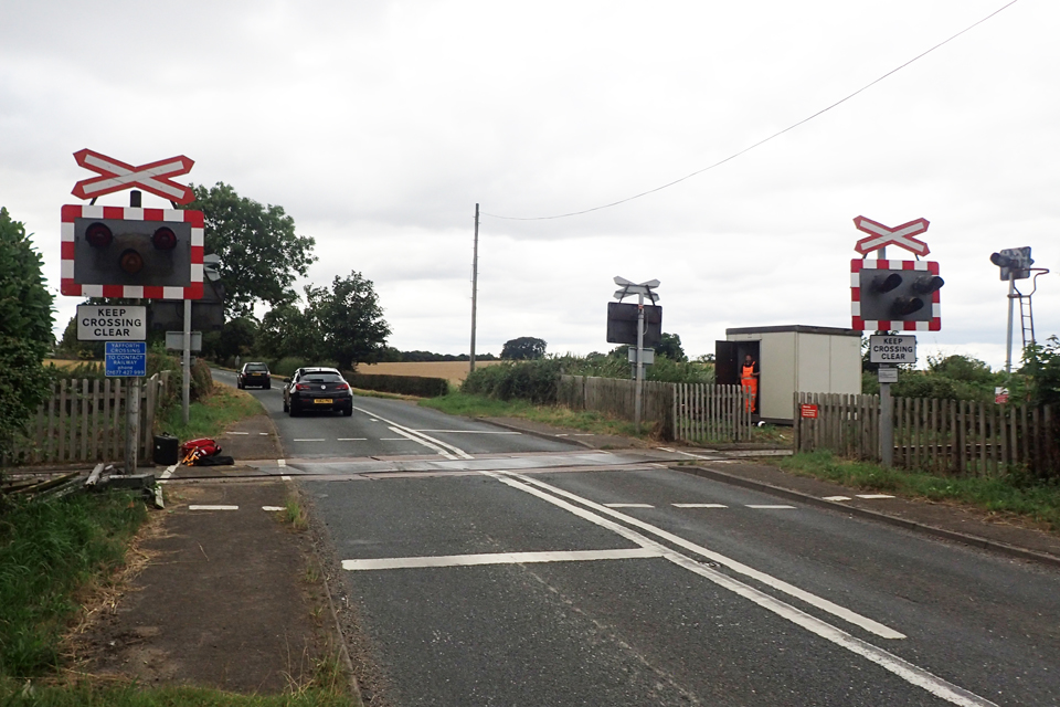Yafforth level crossing on a cloudy day. The traffic warning lights are shown along with warning signs for crossing users. A railway worker standing at the door of a portacabin to the right of the crossing. Two cars are driving away from the camera.
