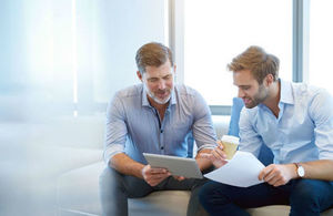 Two male colleagues sit together to chat about work over a mobile tablet device