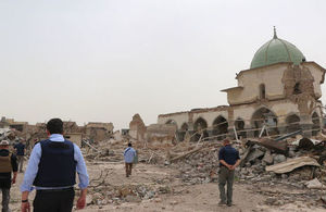 Wide shot of five men in blue shirts, some in flak jackets, walking amidst the rubble of buildings.