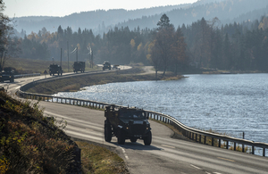 A Foxhound protected patrol vehicle leads a convoy of trucks on a road past a Norwegian lake in the sun.
