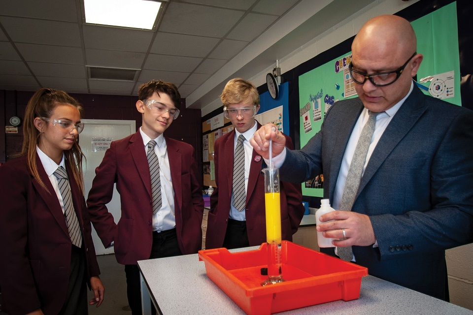 Secondary school pupils wearing safety goggles and watching their teacher pour chemicals into a test tube