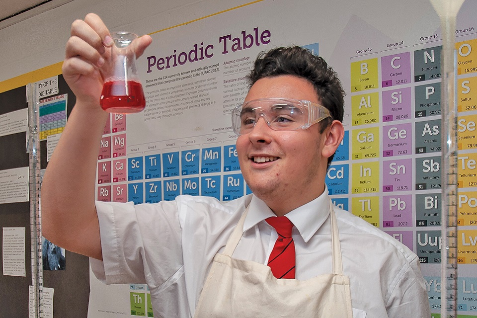 Secondary student wearing safety goggles, standing in front of a poster of the Periodic Table and holding up a beaker with red liquid