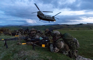 A chinook helicopter flies over a group of Royal Marines wearing camouflage and holding guns during Exercise Joint Warrior 2018