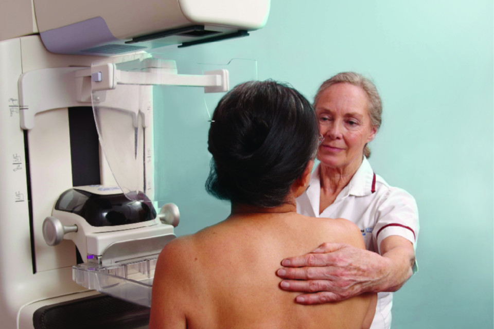 Woman standing next to mammography equipment, being helped into position by a female mammographer. 