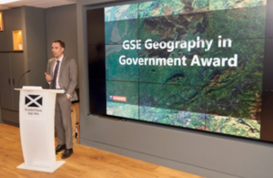 David Wood, GSE Head of Geography, introduces the Geography in Government Awards at Scotland House in London. He is stood behind a podium addressing those in the audience.