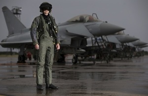 Typhoon pilot in current aircrew equipment assemblies (AEA) stood in front of a row of Typhoon Aircraft with wet pan and some form of water tower in background