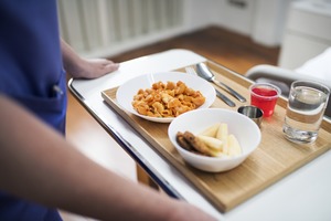 Close-up of a bowl of pasta, a bowl of fruit and a glass of water on a tray being carried by a nurse