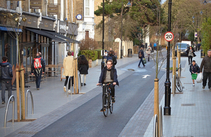 Cyclist on a cycle path passing shops and houses, people walking on pavements. A London mini Holland scheme.