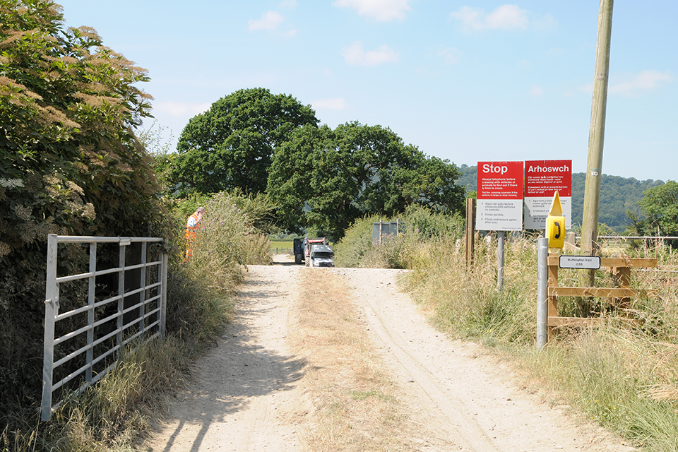 A rural user worked level crossing showing the track across the railway line surrounded by tress. The gate is open. Red and white warning signs are to the right of the track.