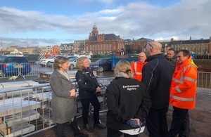 Minister Pow and Emma Howard Boyd stood facing a group of five Environment Agency staff members and local officials next to the Great Yarmouth flood barrier