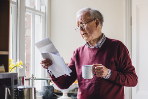 Senior man holding a mug and bills in his hands looking worried.