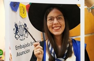 A woman wearing a robe, wizard hat, round glasses and holding a wand smiles at the camera