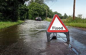 A flood sign is placed on a road. A large flood is in the background with a car driving through the flood water.