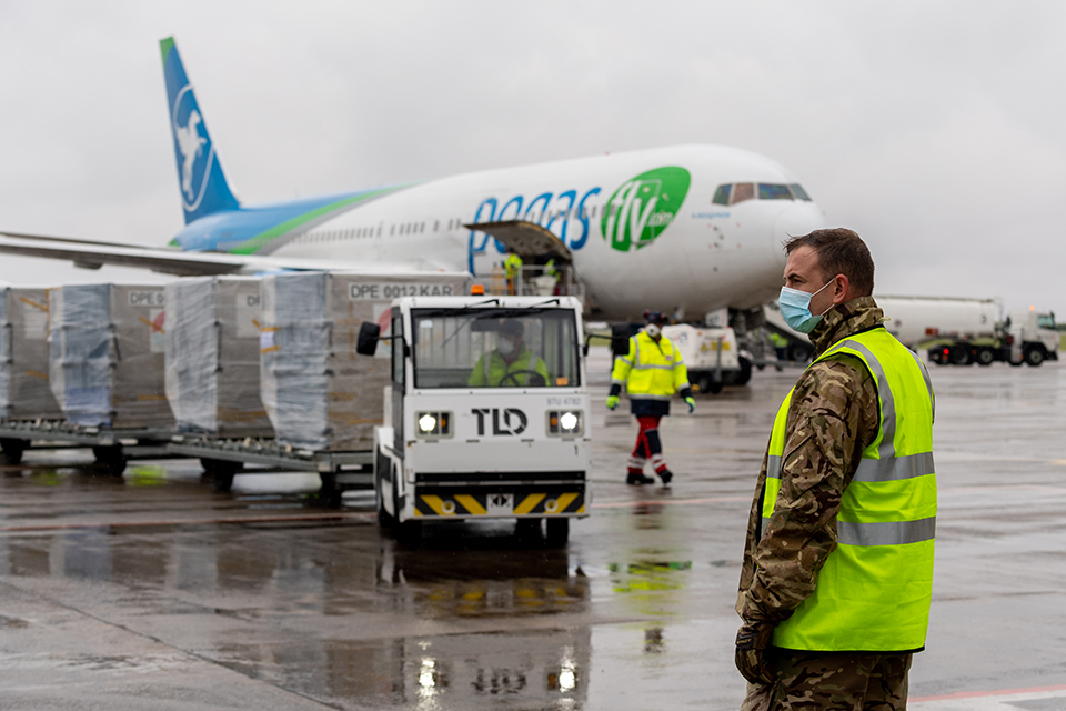 A soldier wearing a facemask stands in an airport. A luggage train loaded with crates of PPE drives towards him.