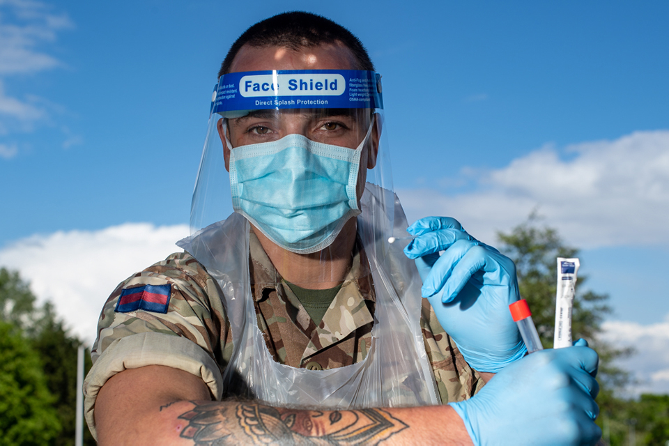 A soldier in Personal Protective Equipment holds a swab out to the viewer.
