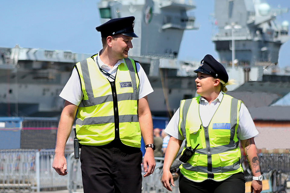 A male and female MOD guard in high visibility jackets walk in front of a ship.