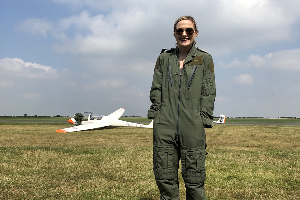 Polly stands smiling in an airfield wearing flight gear with her hands in her pockets and a glider behind her.