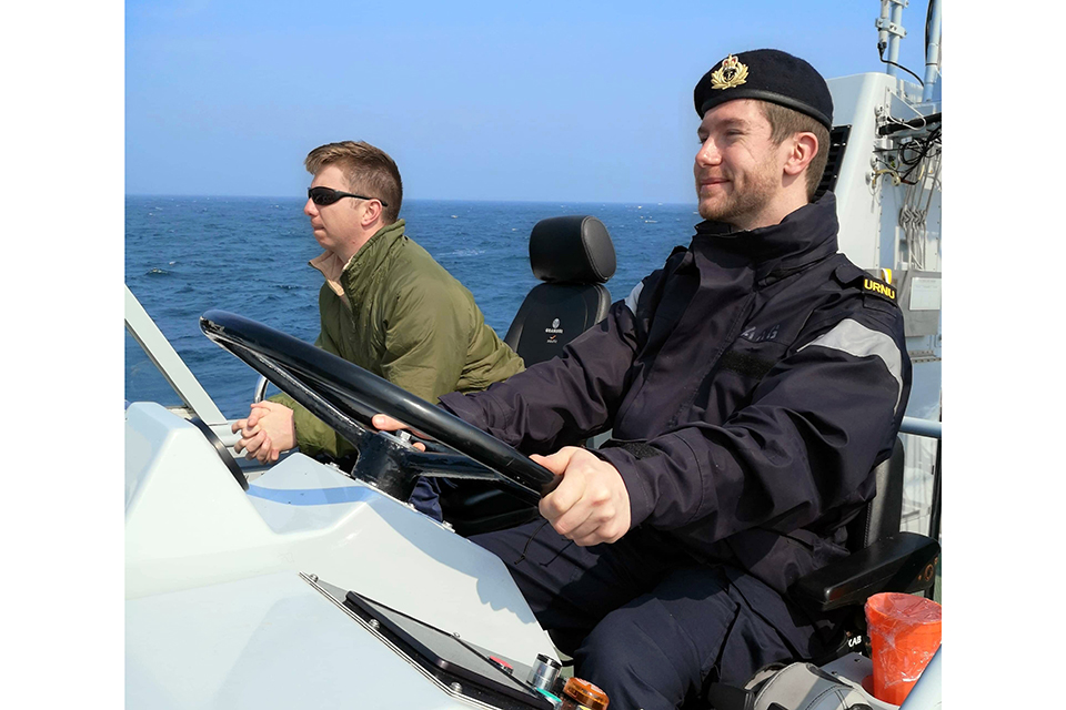 Chris sits smiling at the wheel of a small ship in his Navy uniform. A person in a green coat and sunglasses sits beside him