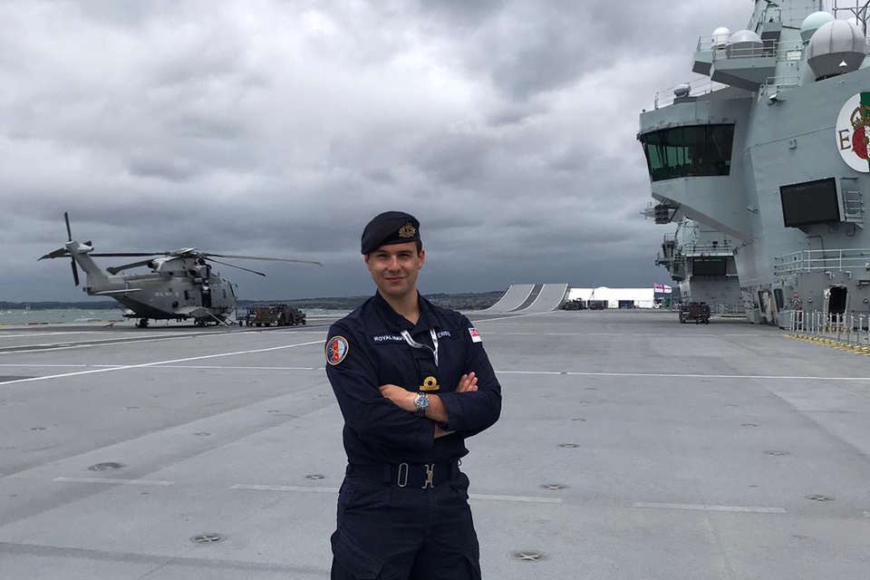 SLT Ewing proudly stands on the grey deck of a ship, with a helicopter, the forward bridge and the ski-jump ramp in the background