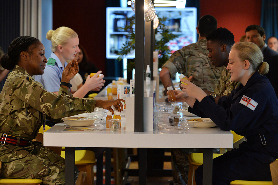 Diners, in military uniform, from all 3 services eat in the new cafe. The cafe looks clean and modern with long white communal tables and yellow seating.