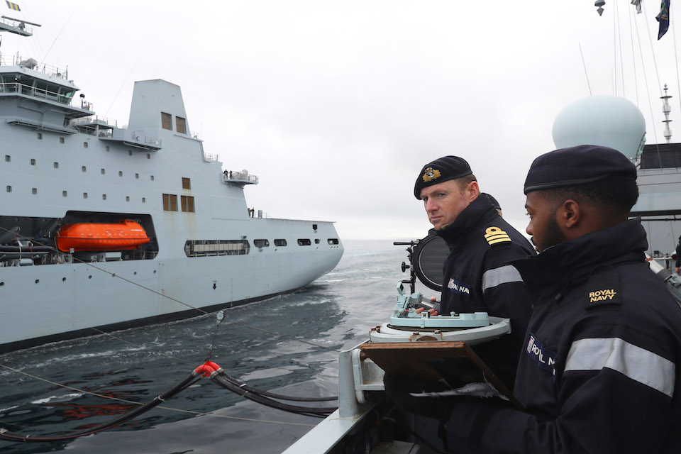 Image depict two Navy personnel on board a ship with another ship in the background.