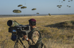 A British Army soldier stands by a weapon in the foreground as paratroopers drop into the field behind him in the background