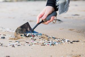 A trowel being held in one hand, collecting plastic debris from the top of a sandy beach